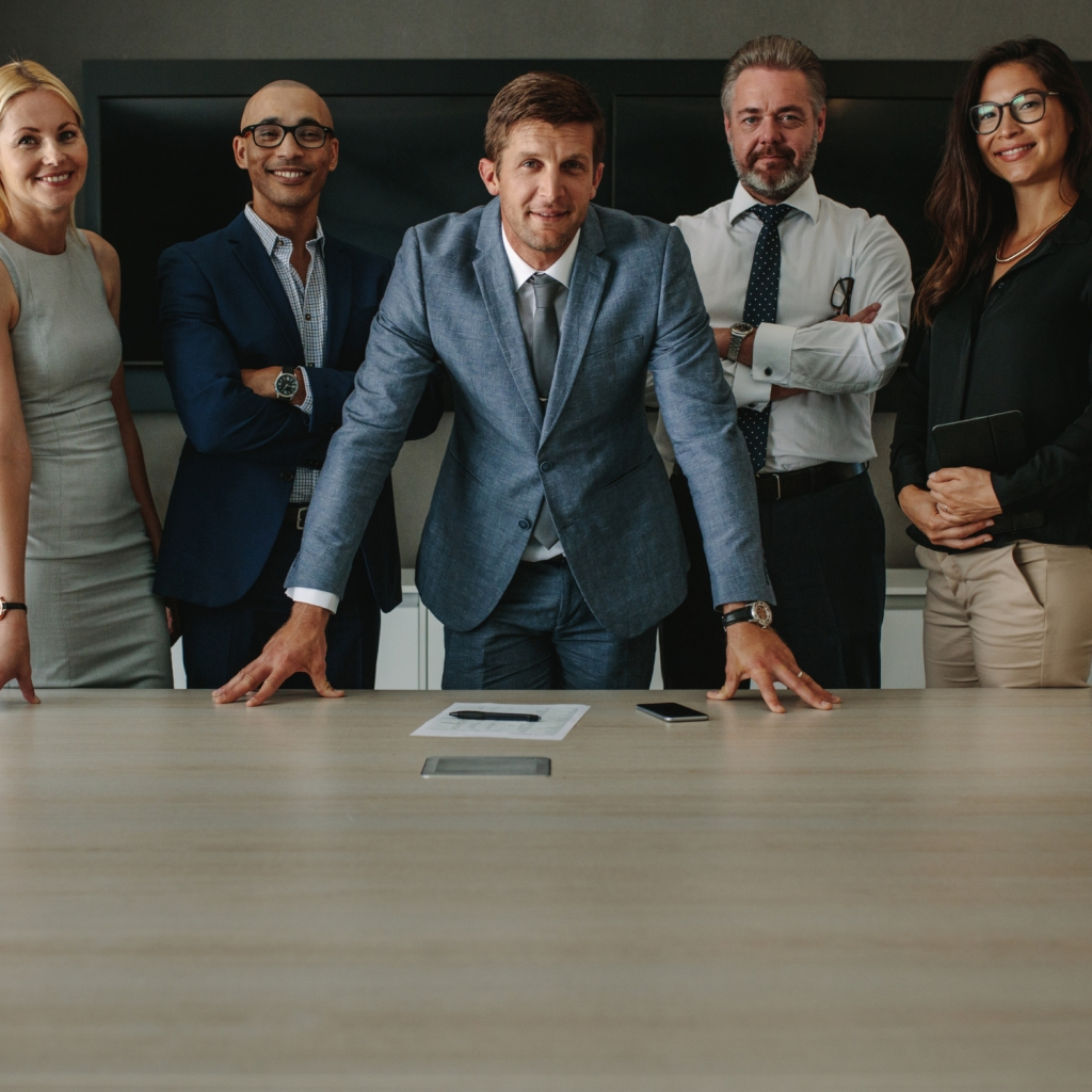 A confident business team standing around a conference table, led by a professional in a blue suit, symbolizing corporate leadership, teamwork, and strategic decision-making.