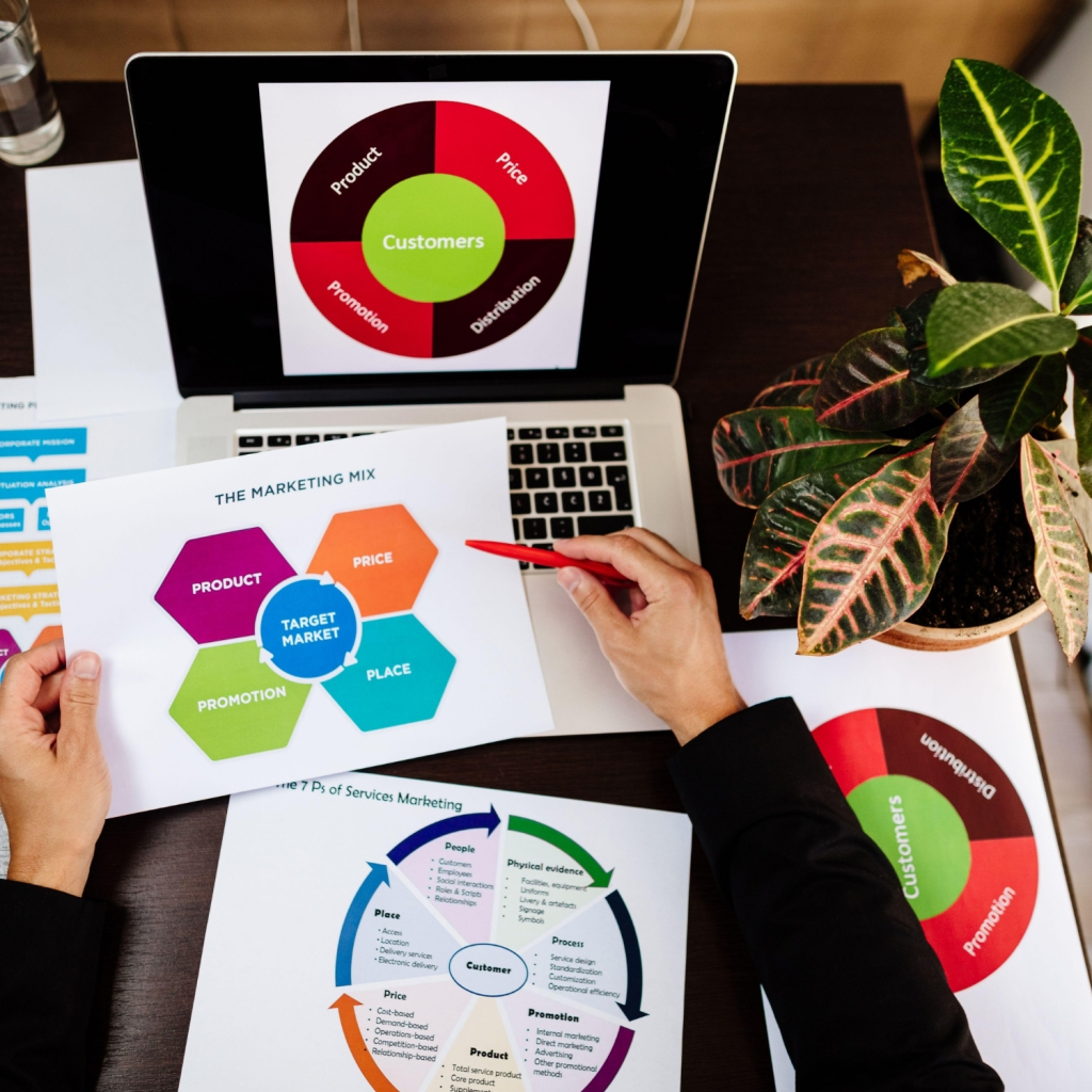 A marketing professional analyzing colorful marketing mix charts on a laptop and printed documents, focusing on customer segmentation, product strategy, and corporate marketing planning.
