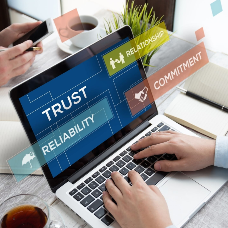 Two people working on laptops and mobile phones at a desk with coffee cups, notebooks, and plants. The laptop screen displays floating digital graphics with the words “Trust,” “Reliability,” “Relationship,” and “Commitment,” symbolizing teamwork, credibility, and long-term professional relationships.