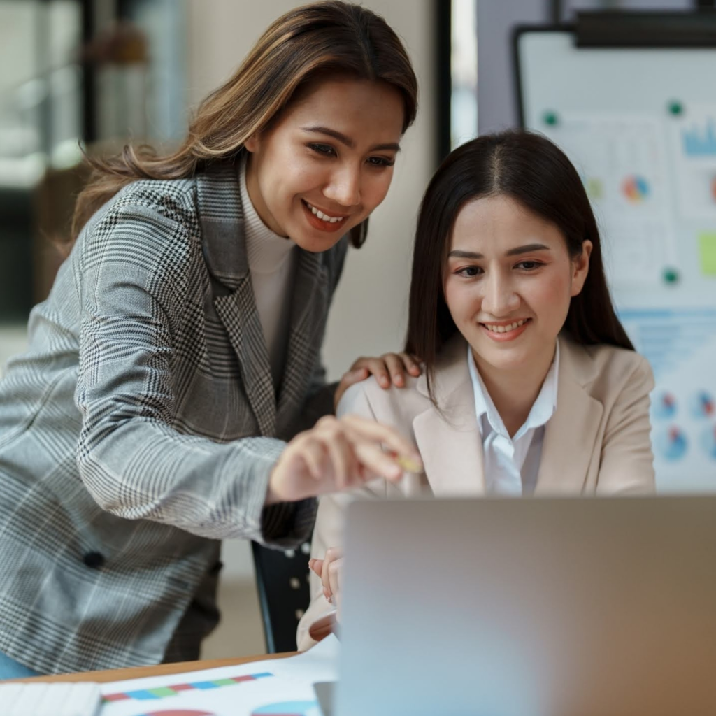 Two professional women collaborating in an office setting, smiling as they look at a laptop screen together. One gestures toward the screen while the other listens attentively, symbolizing teamwork, mentorship, and positive workplace collaboration.