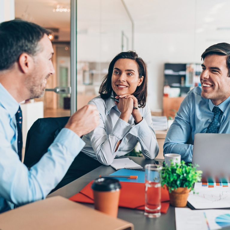 A group of business professionals engaged in a meeting, smiling and discussing ideas around a modern office table with laptops, documents, and coffee.