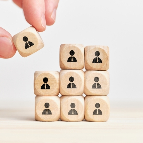 This image depicts a hand placing a wooden cube with a human figure icon on top of a stack of other cubes, each displaying the same icon. The stack of cubes is arranged in a pyramid shape, symbolizing growth, team-building, or leadership. The simple wooden cubes and minimalist design convey a sense of organization, collaboration, and progress in a professional or business context.