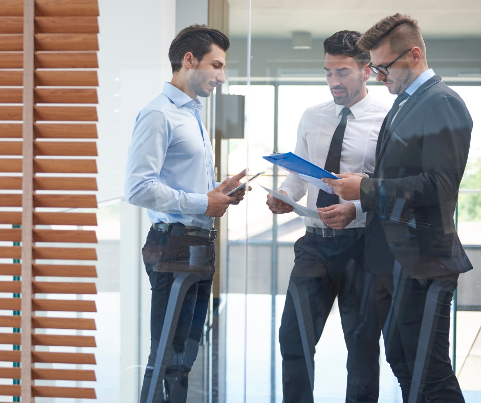 Three business professionals discussing strategy documents in a modern office environment, representing collaborative B2B decision-making, enterprise planning, stakeholder alignment, and data-driven business operations.