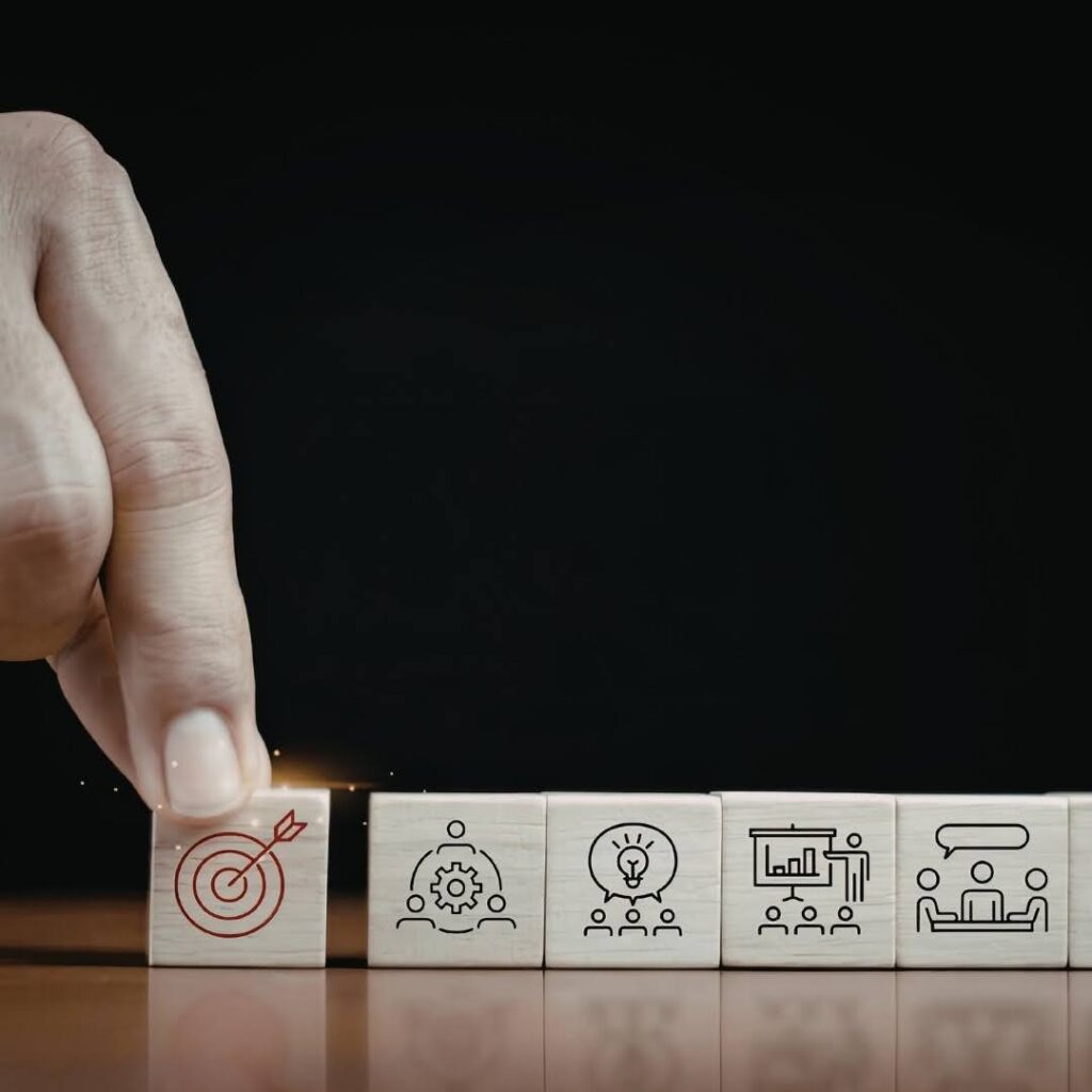 A strategic business planning and performance management visual featuring a hand placing a target icon block alongside symbols representing collaboration, innovation, analytics and corporate communication against a dark background.