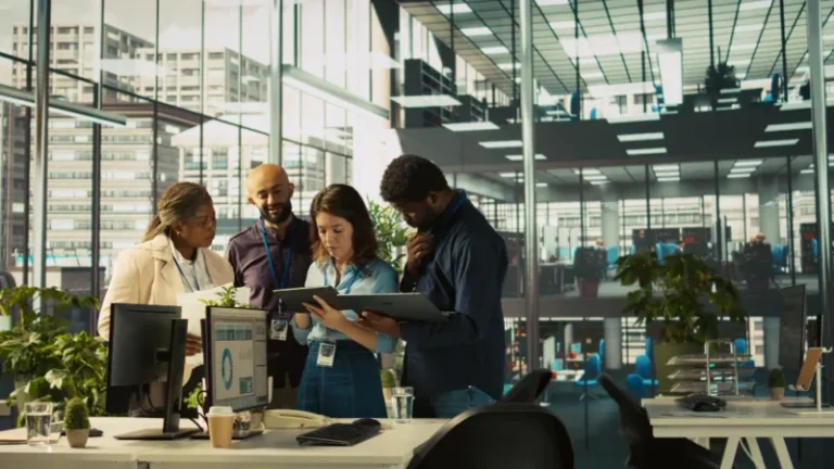 A diverse B2B marketing team collaborates around laptops and digital dashboards in a modern office, reviewing data, strategy and campaign performance.